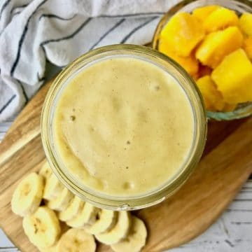 A yellow smoothie next to banana slices and mango chunks.